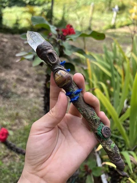 Una mano sostiene una varita de poder de porcelanicrón con una punta de cuarzo, frente a un jardín